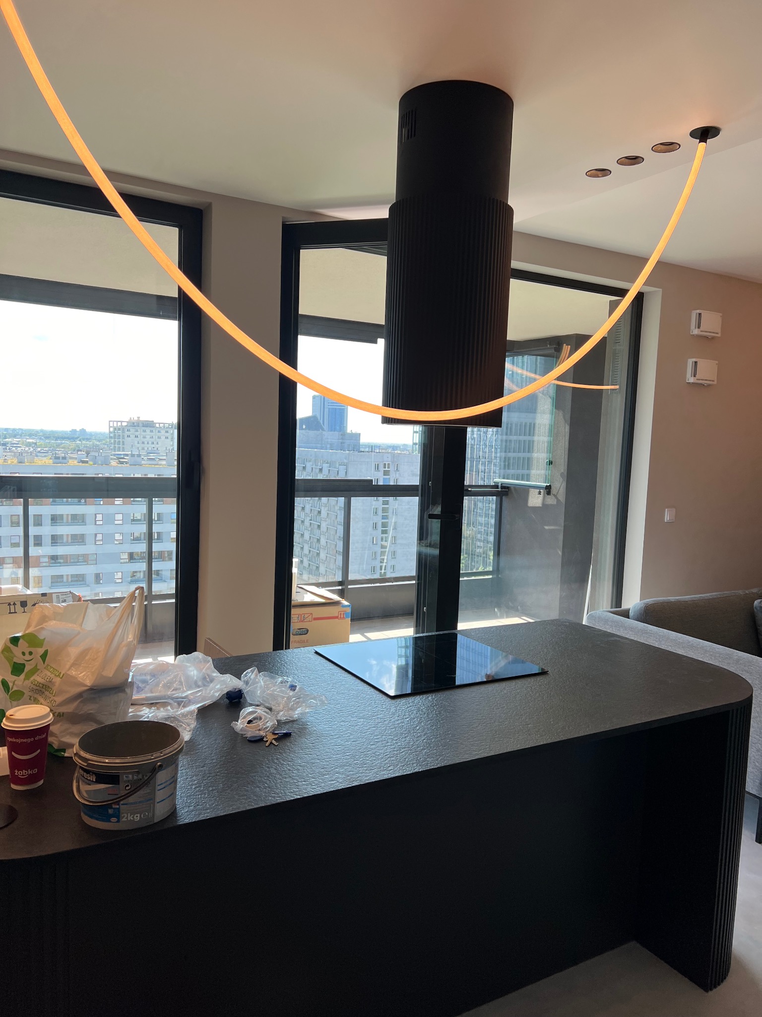 Nowoczesna kuchnia z wyspą, czarnym okapem cylindrycznym i listwą LED pod sufitem; na blacie wiadro kleju, kubek po kawie i folie ochronne.