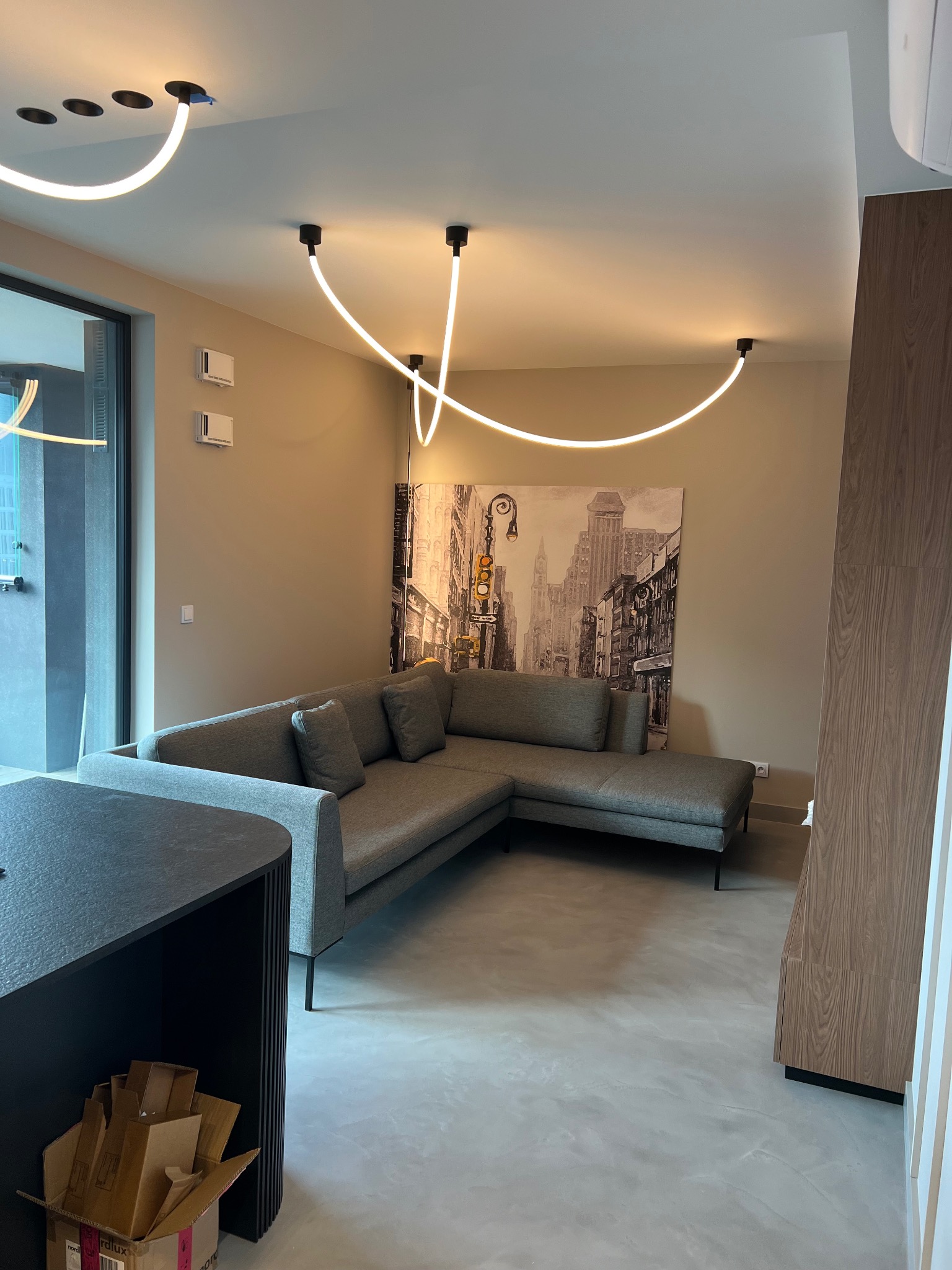 Nowoczesny salon z szarą sofą narożną, betonową podłogą, obrazem z motywem miejskim i nietypowym oświetleniem sufitowym w postaci białych, zakrzywionych lamp LED.