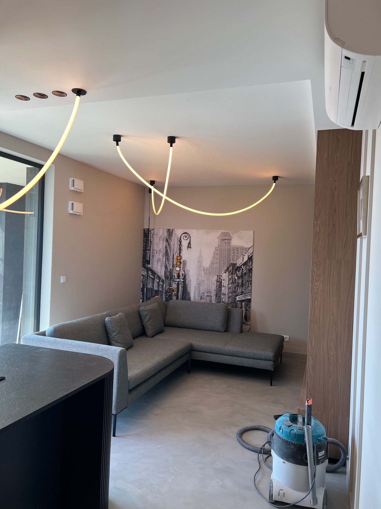 Nowoczesny salon w neutralnych kolorach z szarą sofą narożną, abstrakcyjnym oświetleniem sufitowym i odkurzaczem przemysłowym stojącym w rogu pokoju.