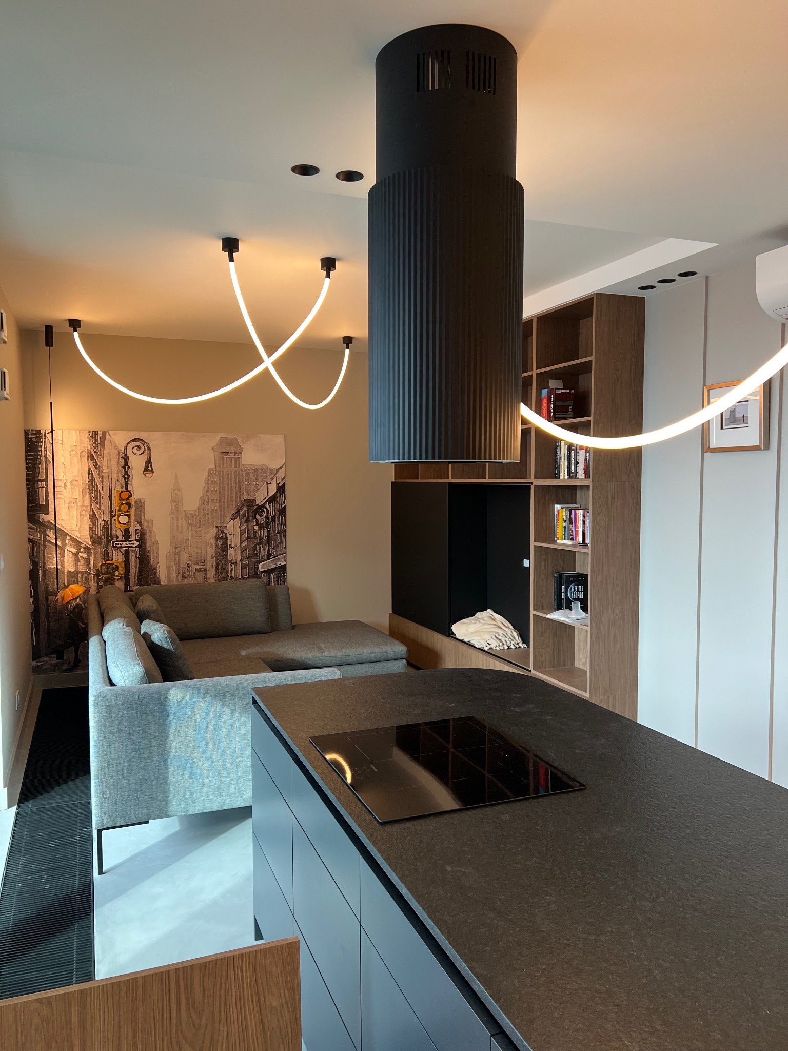 Nowoczesne wnętrze z szarą sofą, czarnym blatem kuchennym z płytą indukcyjną, designerskim oświetleniem neonowym oraz regałem z książkami w tle.