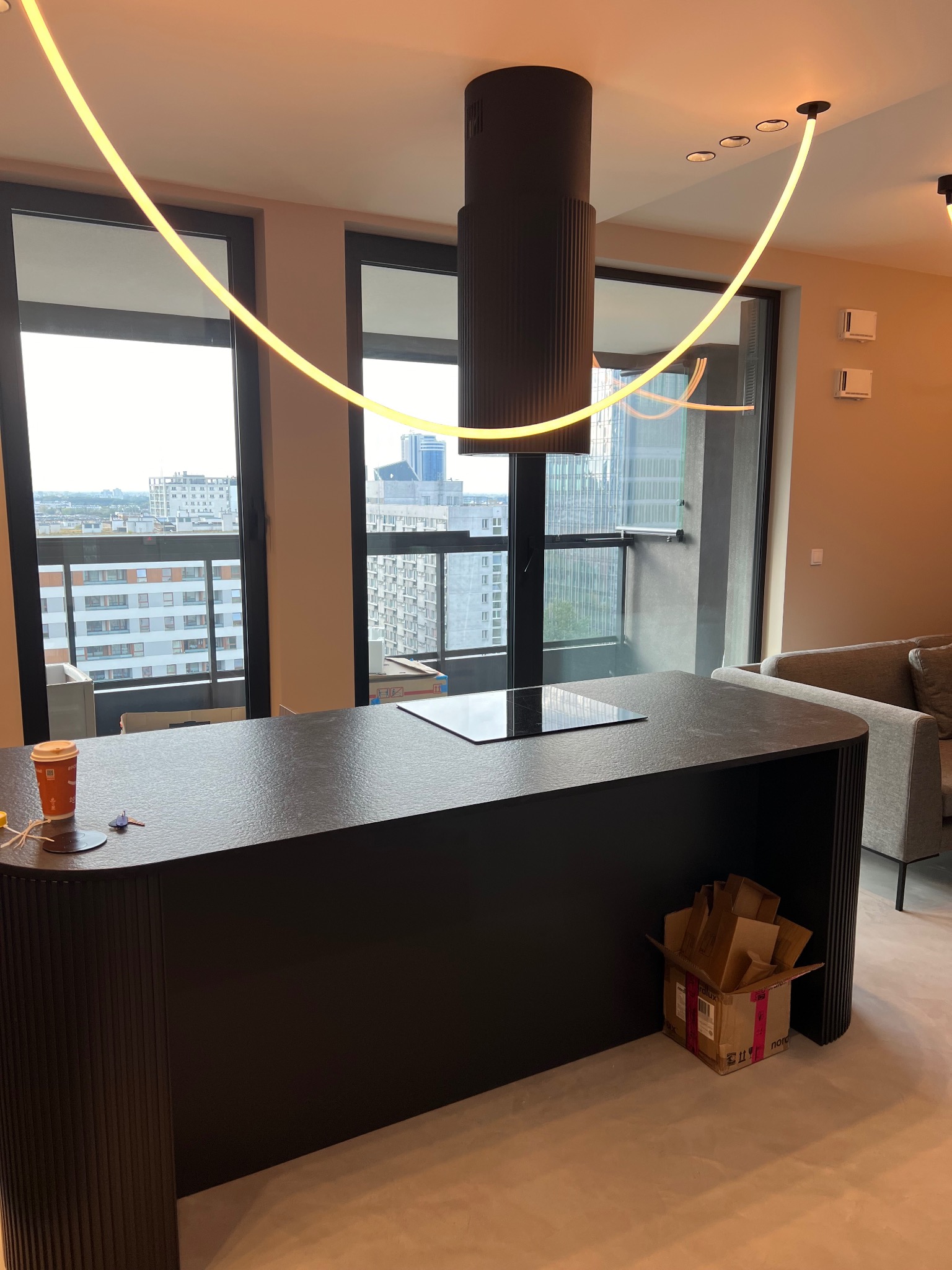 Nowoczesna kuchnia z wyspą o czarnym, teksturowanym blacie, cylindrycznym okapem i futurystycznym oświetleniem LED, widok na miasto za oknem, karton pod wyspą.