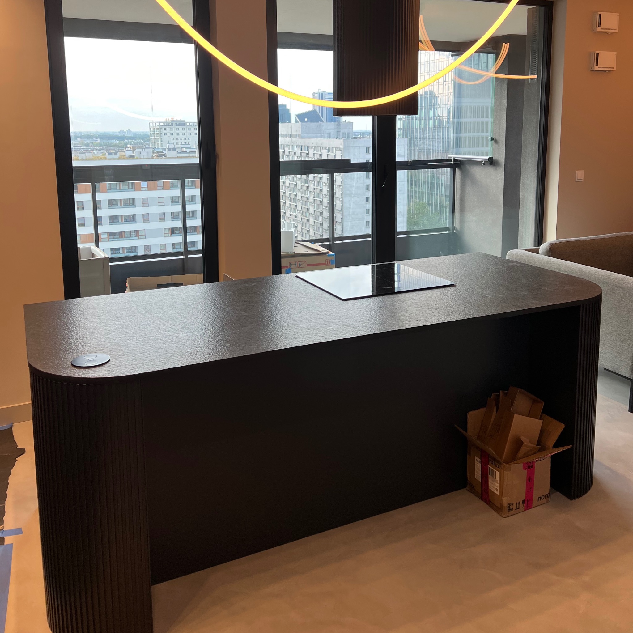 Czarny, teksturowany blat kuchenny z płytą indukcyjną, widok na miasto przez okno, kartonowe pudełko u dołu, nowoczesne oświetlenie sufitowe.