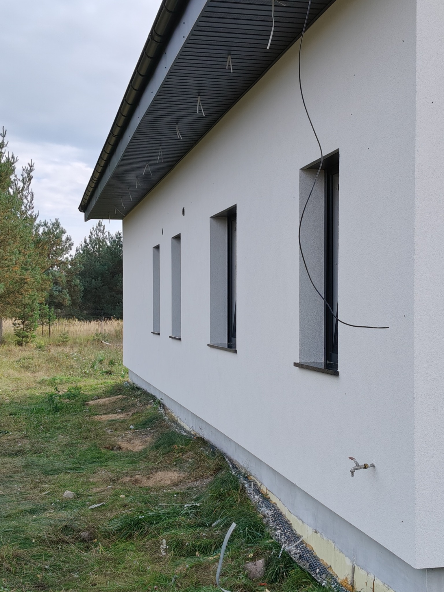 Elewacja domu jednorodzinnego z oknami w czarnych ramach, widok z boku, trawa i drzewa w tle. Widoczne wystające kable i kran na ścianie.