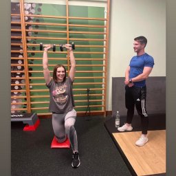 Szymon Adamowicz Trener Osobisty - Kobieta w szarej koszulce z nadrukiem wykonuje wykrok z hantlami nad głową na czerwonej macie, mężczyzna w niebieskiej koszulce obserwuje, w tle drabinki gimnastyczne i butelka wody.