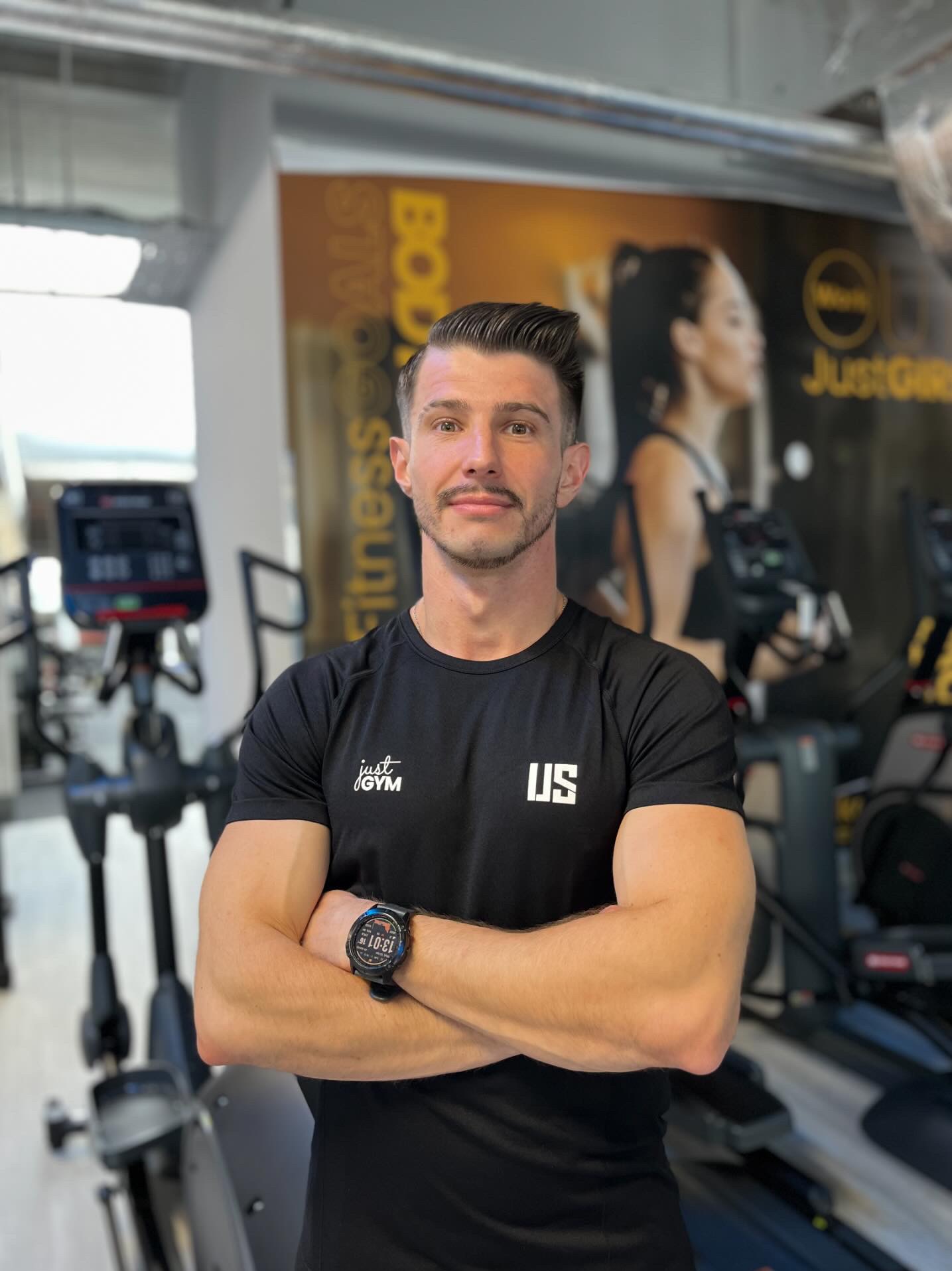 Mężczyzna z wąsami, ubrany w czarną koszulkę z logo 'Just Gym' i zegarek sportowy na nadgarstku, stoi na tle siłowni z maszynami do ćwiczeń i plakatem reklamowym.