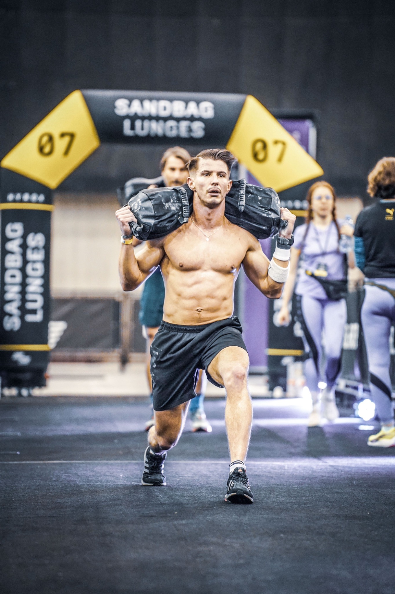 Mężczyzna w Lublinie wykonuje wykroki z obciążeniem (workiem z piaskiem) na ramionach podczas treningu funkcjonalnego na tle bramki z napisem 'Sandbag Lunges'.
