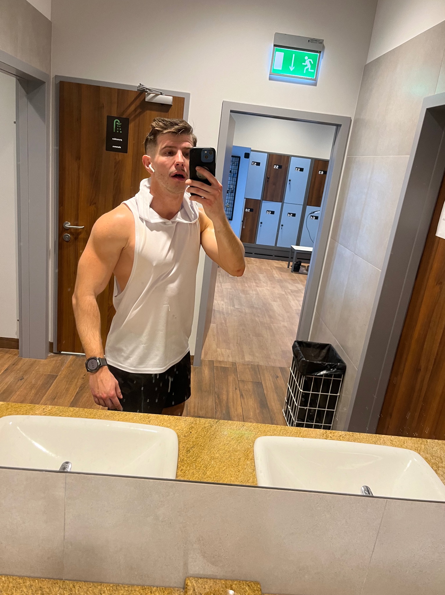 Mężczyzna w sportowym stroju robi selfie w szatni siłowni, widoczne szafki, umywalki i znak ewakuacyjny.