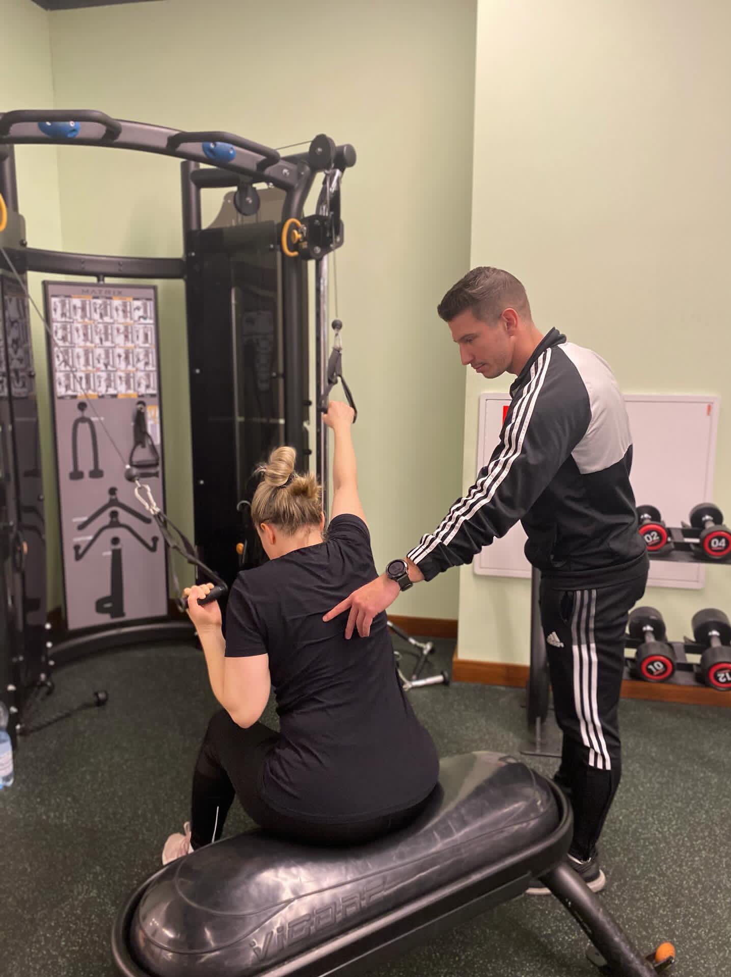 Kobieta ćwiczy na maszynie pod okiem mężczyzny w sportowym stroju w siłowni, mężczyzna koryguje jej postawę, w tle widać hantle i tablicę.