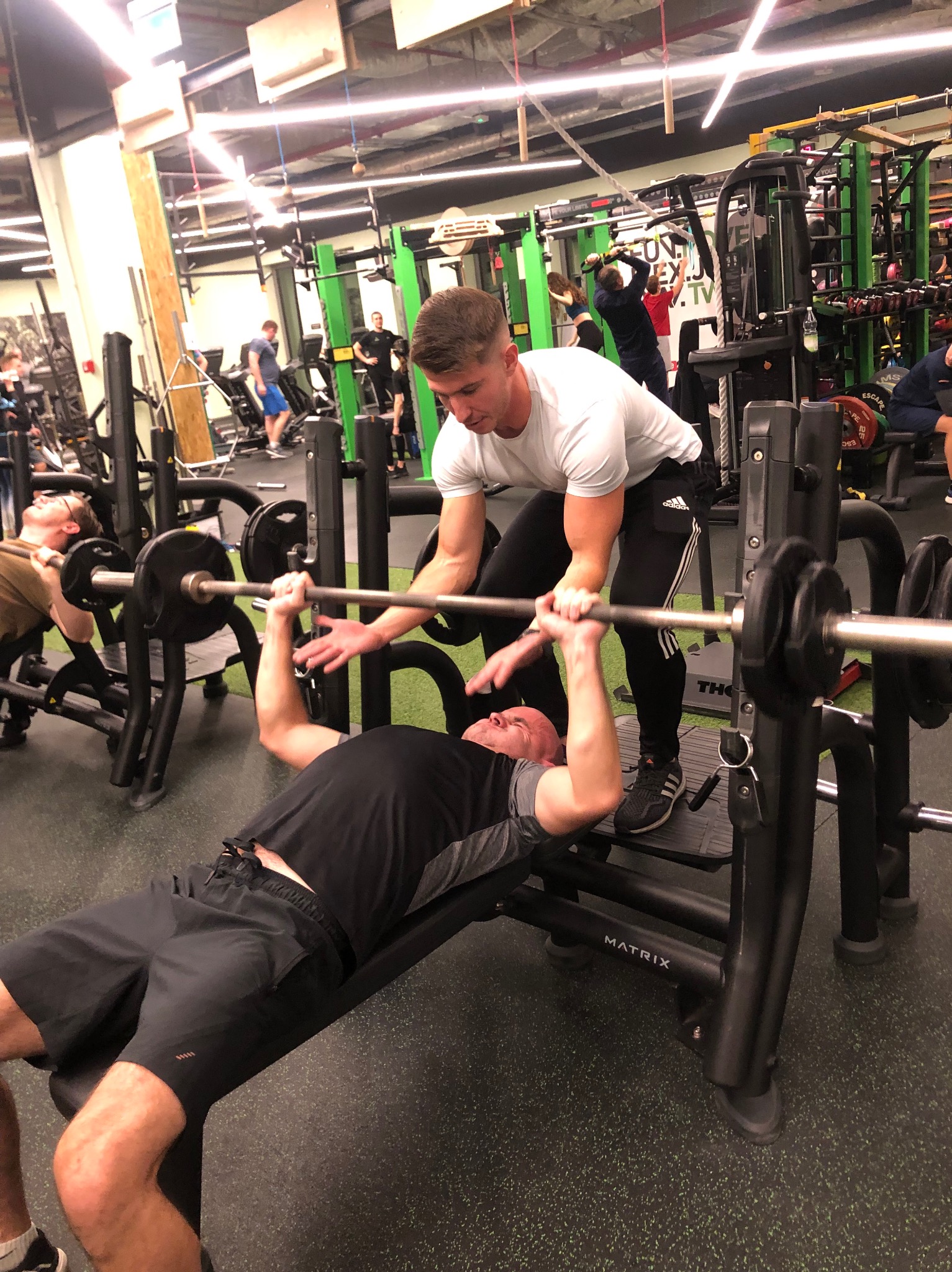 Mężczyzna ćwiczy wyciskanie sztangi na ławce pod okiem instruktora w nowoczesnej siłowni z innymi osobami w tle.