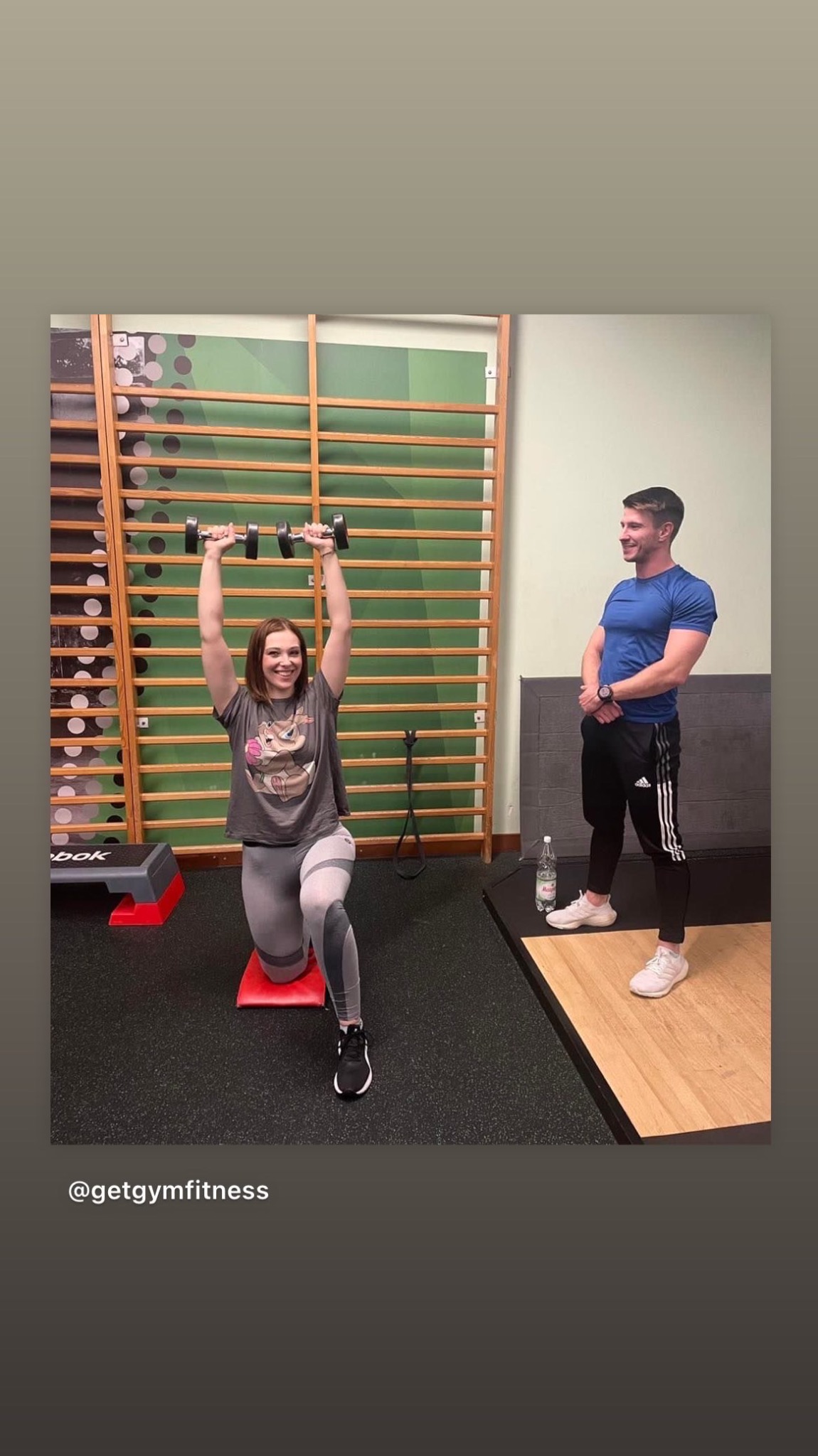 Kobieta w szarej koszulce z nadrukiem wykonuje wykrok z hantlami nad głową na czerwonej macie, mężczyzna w niebieskiej koszulce obserwuje, w tle drabinki gimnastyczne i butelka wody.