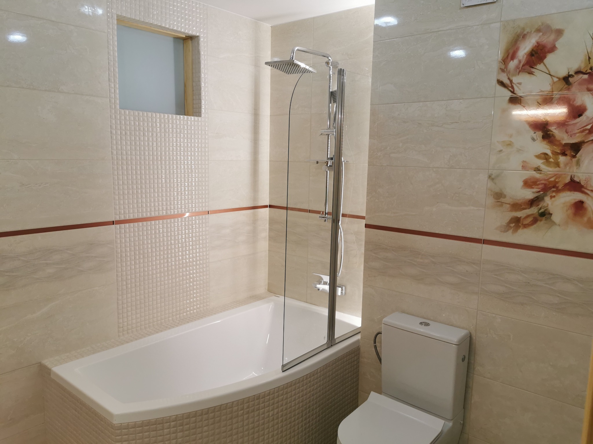 Jasna łazienka z beżowymi płytkami, kabiną prysznicową nad wanną, toaletą i dekoracyjną płytką z motywem kwiatowym.