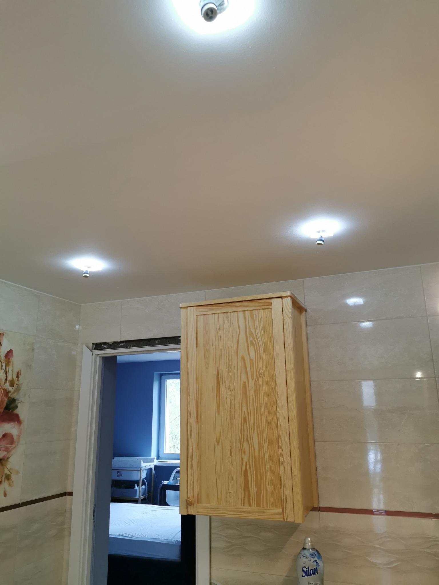 Wykończona łazienka z beżowymi płytkami, drewnianą szafką podwieszaną i widokiem na pokój przez otwarte drzwi.