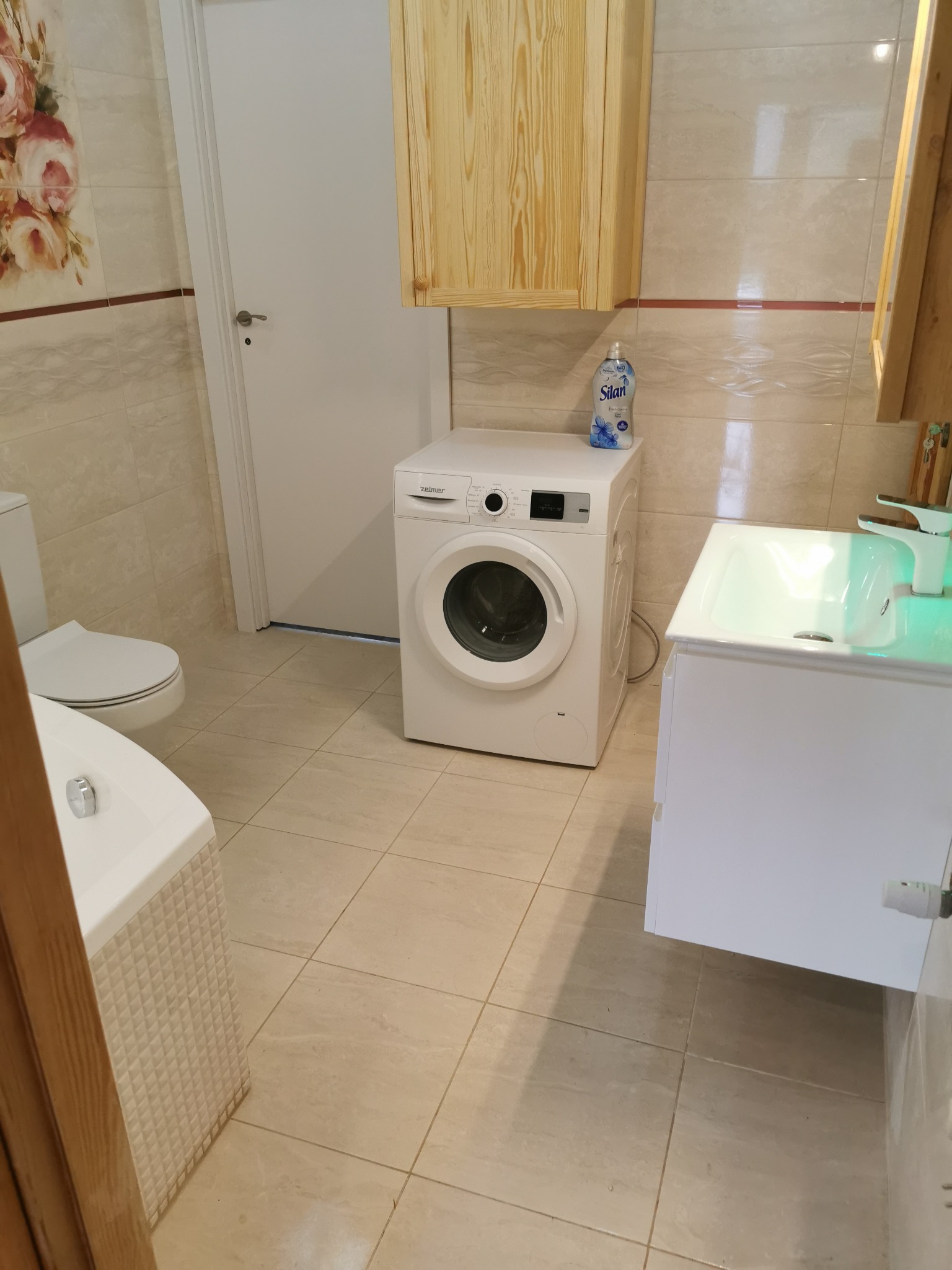 Jasna łazienka w beżowych płytkach z pralką, umywalką z szafką i toaletą. Na pralce stoi butelka płynu do płukania Silan. Nad pralką wisi drewniana szafka.