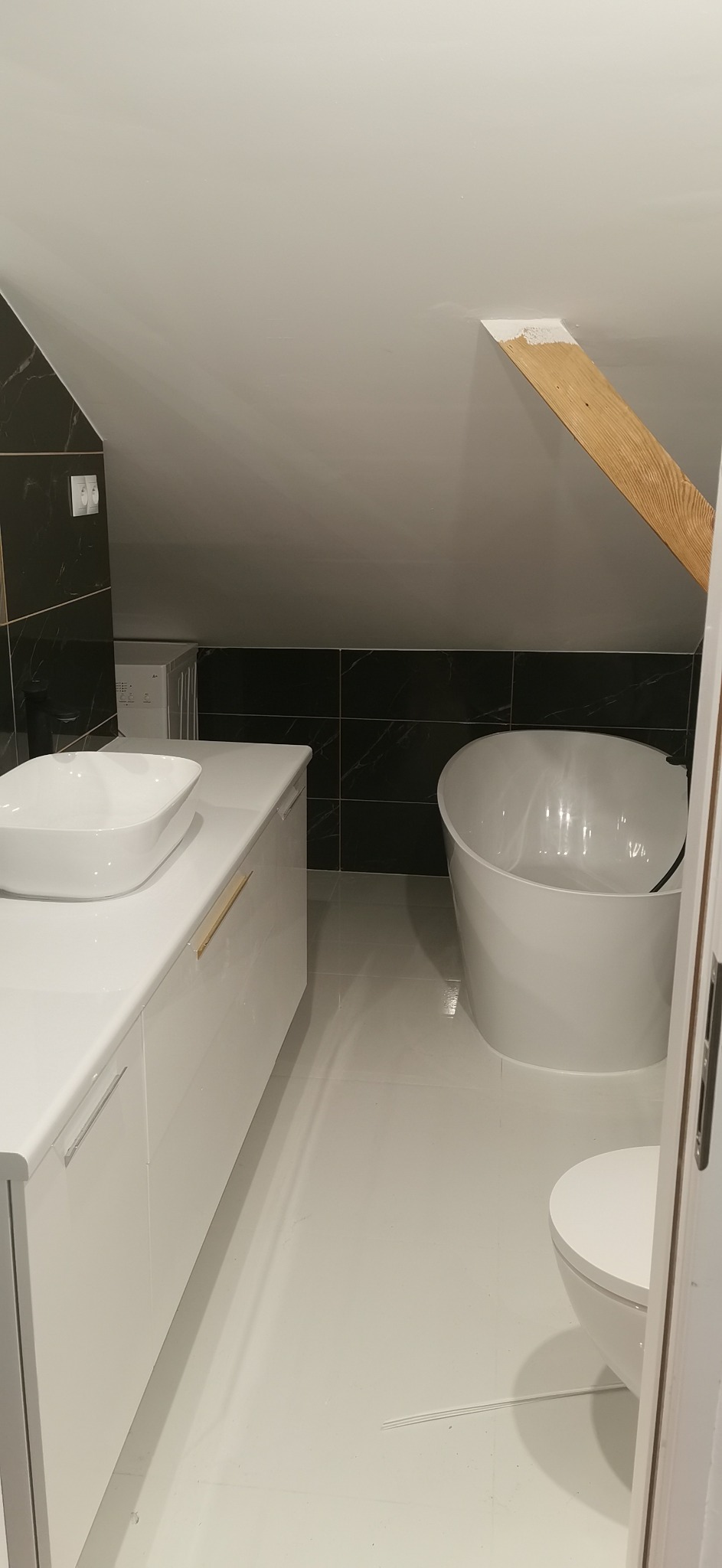 Nowoczesna łazienka na poddaszu z białą armaturą: wolnostojąca wanna, umywalka nablatowa na szafce z minimalistycznymi uchwytami, czarne płytki imitujące marmur na ścianie, drewniany element...