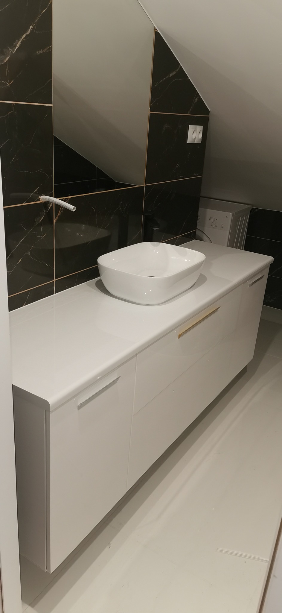 Nowoczesna łazienka na poddaszu z białą umywalką nablatową o kwadratowym kształcie, szafką pod umywalką z połyskującymi białymi frontami i złotymi uchwytami, czarnymi płytkami imitującymi marmur...