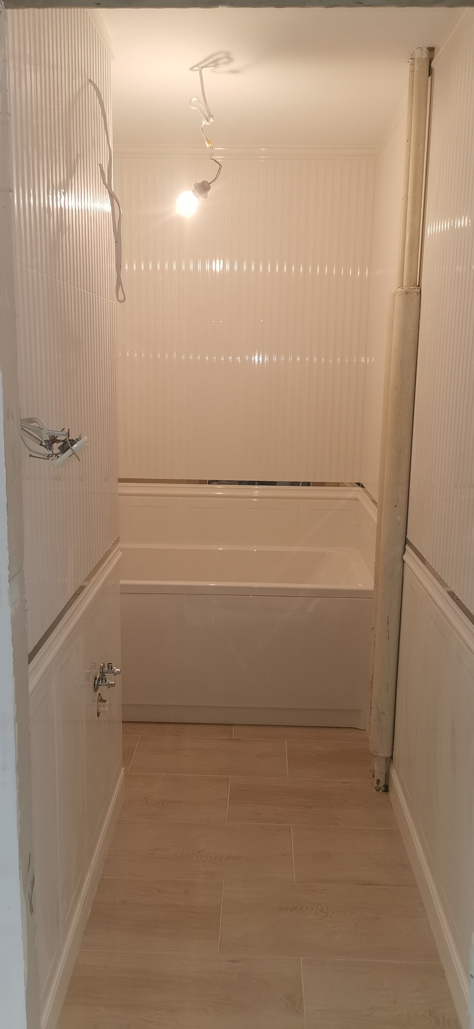 Wnętrze łazienki w trakcie remontu: widoczne płytki ścienne z pionowymi rowkami, jasna podłoga z płytek imitujących drewno, wanna, rury oraz wisząca żarówka bez oprawy. Częściowo wykończone ściany...