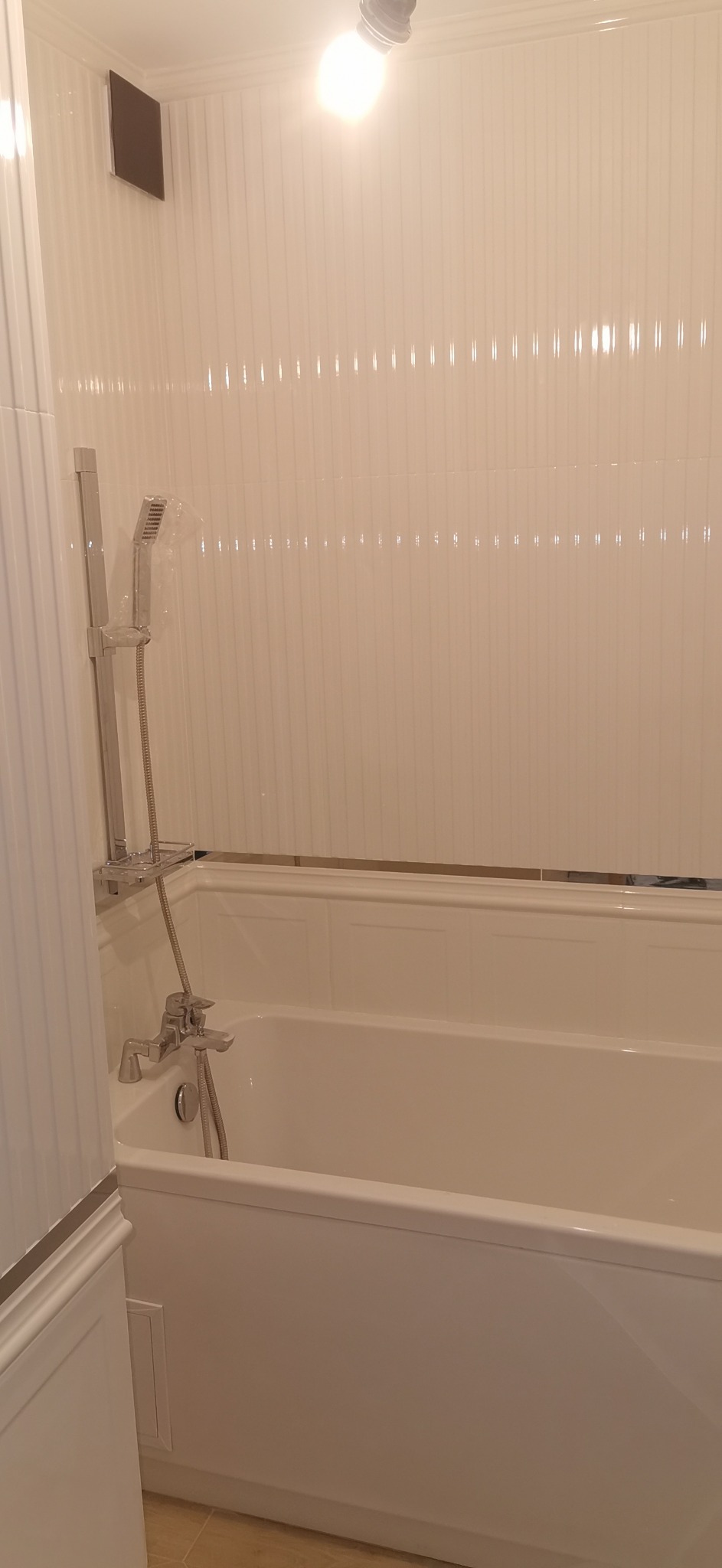 Biała wanna z chromowaną baterią i panelem prysznicowym na tle białych, pionowo żłobkowanych paneli ściennych i sufitu z halogenem.