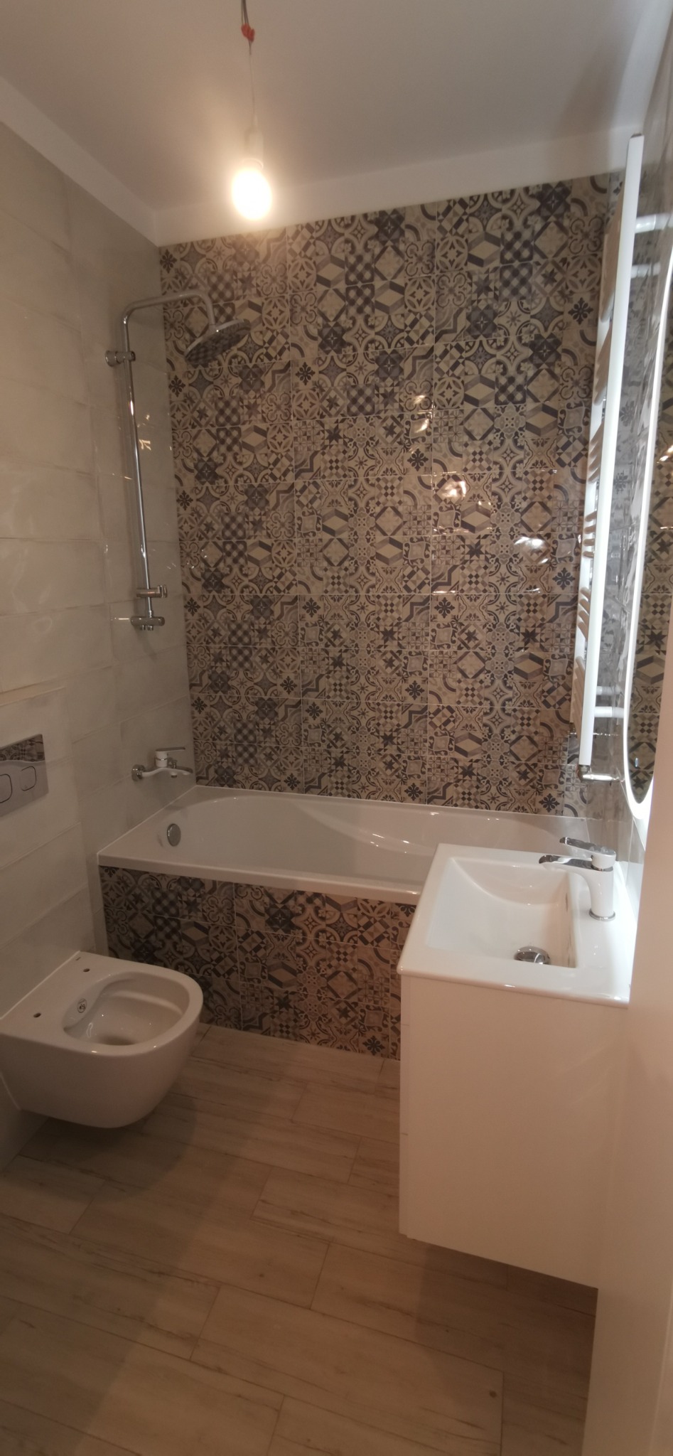 Wykończona łazienka z wzorzystymi płytkami na ścianie przy wannie, wisząca toaleta, umywalka z szafką i drewnopodobna podłoga.