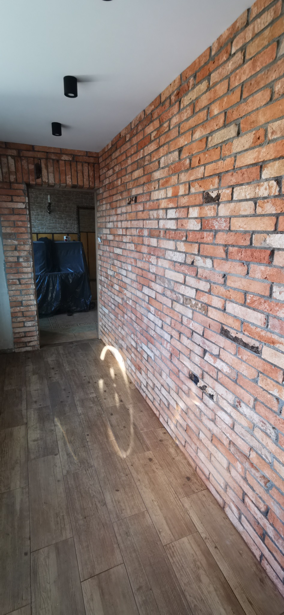 Pomieszczenie po remoncie z ceglaną ścianą, drewnopodobną podłogą i widocznym przejściem do kolejnego pomieszczenia, w którym meble są przykryte folią ochronną.