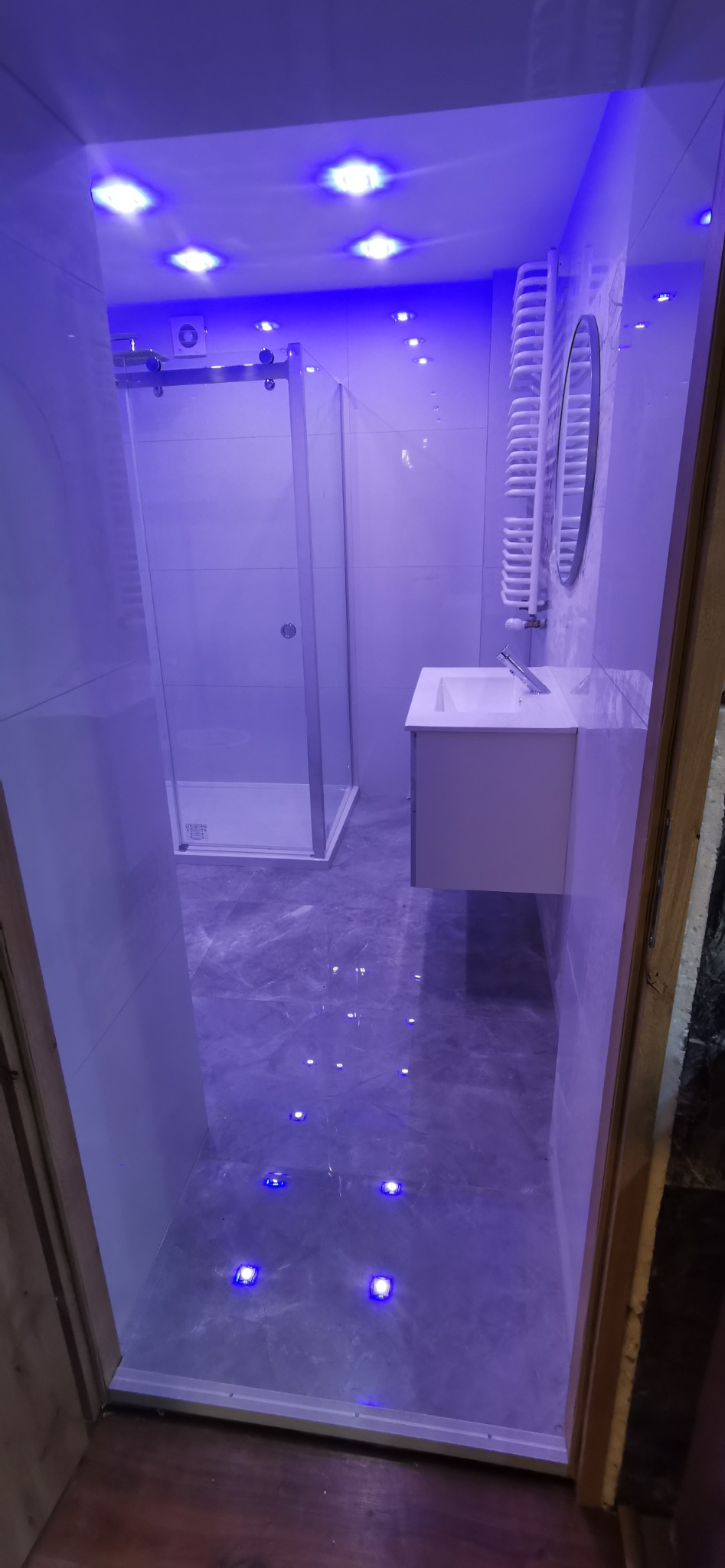 Nowoczesna łazienka z szarą marmurową podłogą, kabiną prysznicową ze szklanymi drzwiami przesuwnymi, białą umywalką z szafką i okrągłym lustrem, oświetlona niebieskim światłem z sufitu i podłogi.