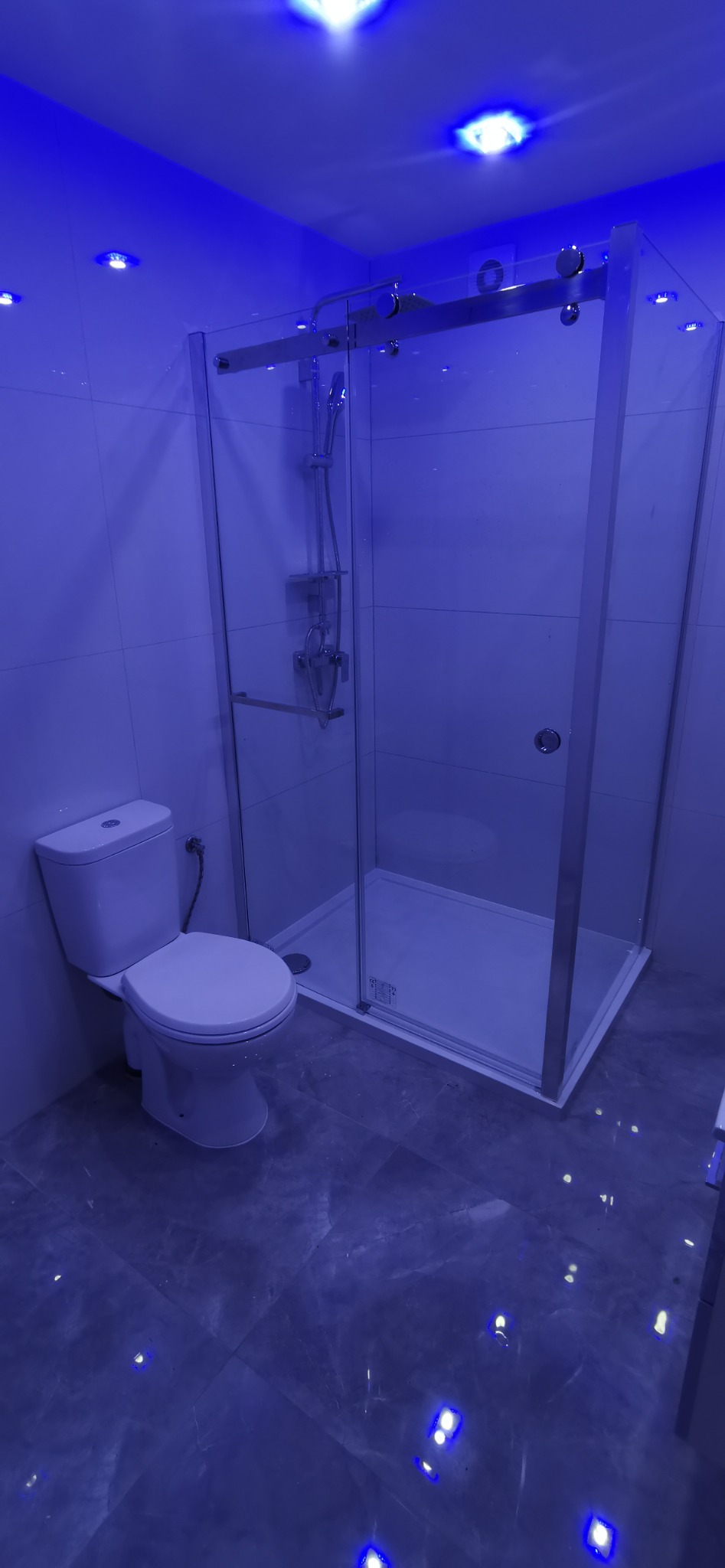 Nowoczesna łazienka z kabiną prysznicową i toaletą, oświetlona niebieskim światłem LED, ukazująca marmurkową posadzkę i białe płytki ścienne.