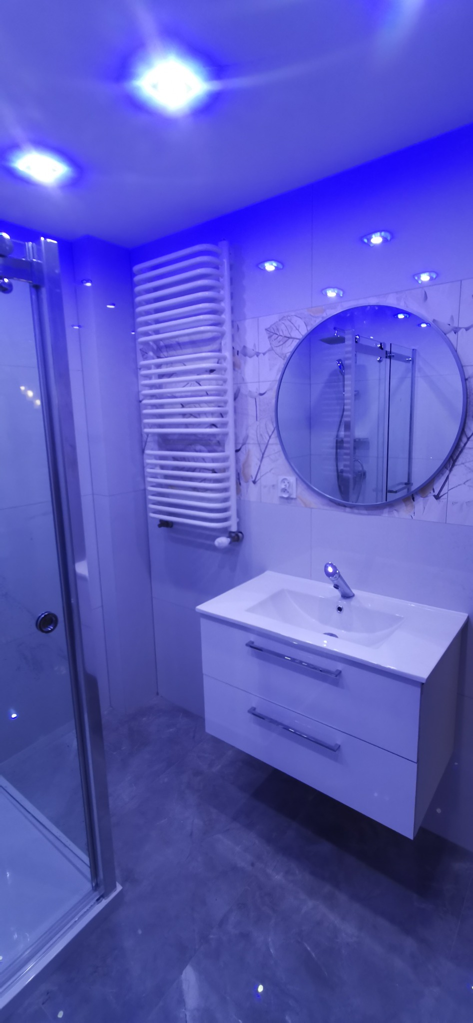 Nowoczesna łazienka z kabiną prysznicową, białą szafką z umywalką i okrągłym lustrem, oświetlona niebieskim światłem LED.