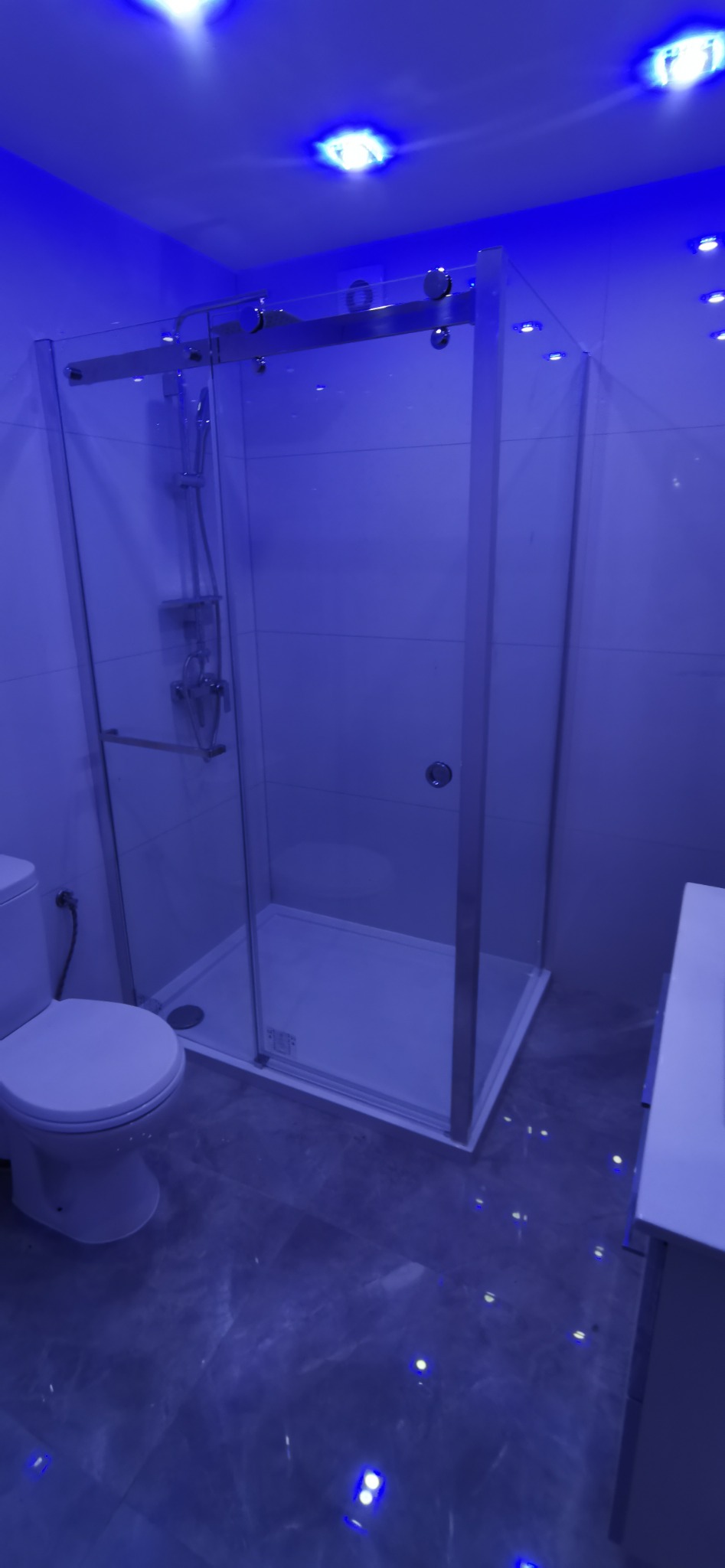 Nowoczesna kabina prysznicowa z przesuwnymi drzwiami ze szkła hartowanego, oświetlona niebieskimi diodami LED w suficie, widoczna toaleta i fragment umywalki.