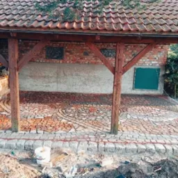 Zadaszone patio w trakcie budowy z wzorem z czerwonej kostki brukowej ułożonej w okręgi, obrzeża z szarej kostki, drewniana konstrukcja dachu, ściana z cegły w tle.