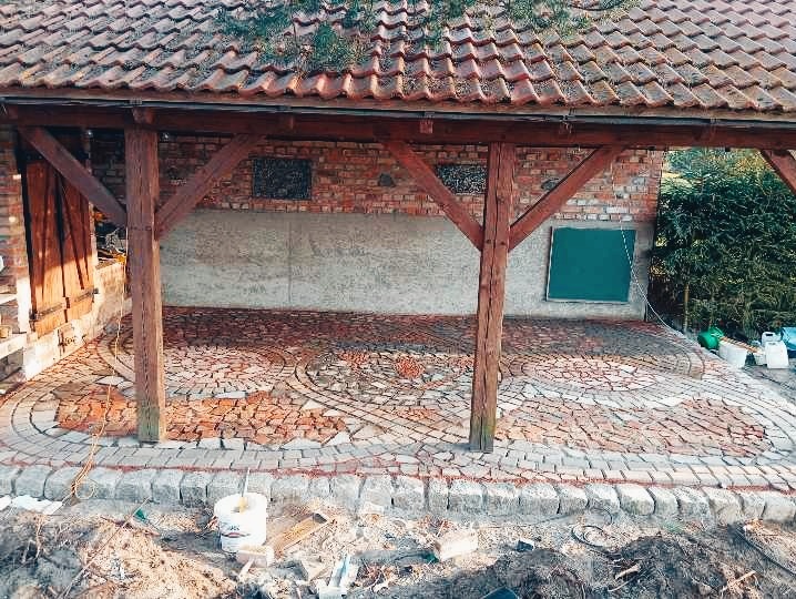 Zadaszone patio w trakcie budowy z wzorem z czerwonej kostki brukowej ułożonej w okręgi, obrzeża z szarej kostki, drewniana konstrukcja dachu, ściana z cegły w tle.