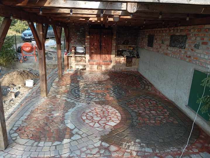 Zadaszony taras z mozaikową nawierzchnią z kostki brukowej w odcieniach czerwieni i szarości, z elementami dekoracyjnymi w kształcie okręgów. W tle widoczna murowana ściana z cegły i drewniane...