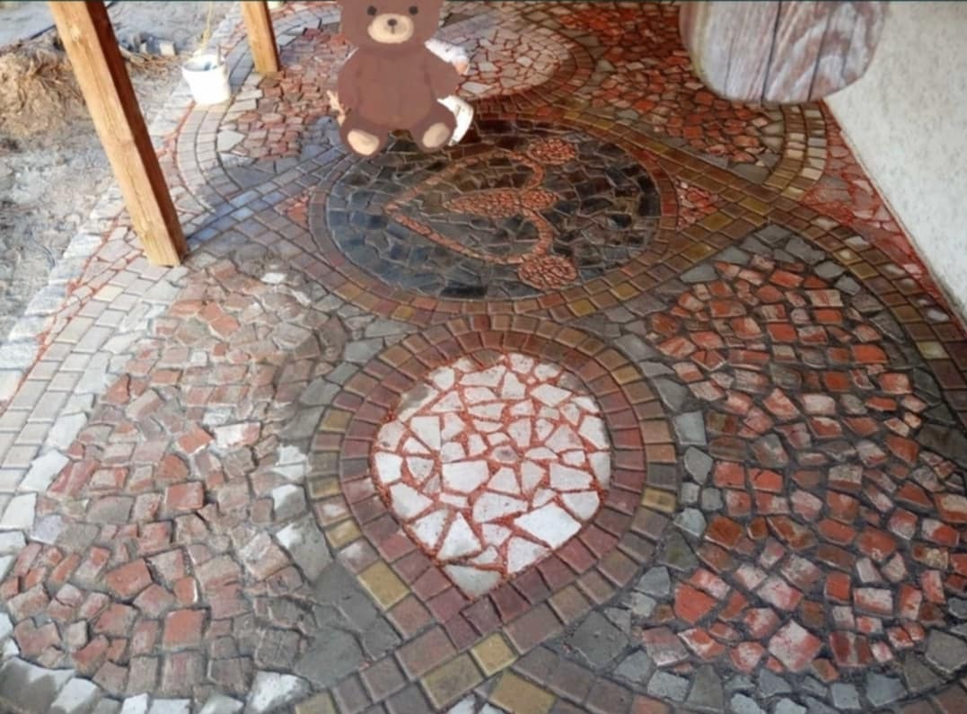 Mozaika z kostki brukowej tworząca wzory, w tym okrągłe motywy i ornamenty, z dodatkiem kartonowego misia dla skali i humoru.