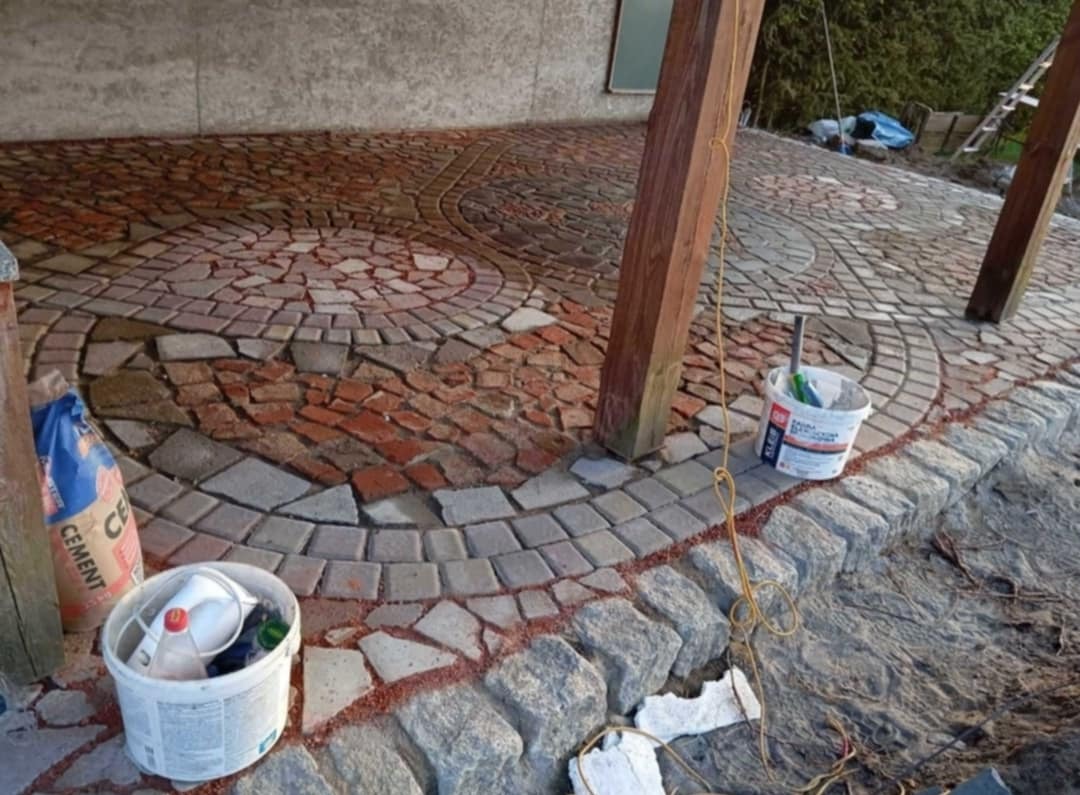 Niedokończona powierzchnia z kostki granitowej i brukowej ułożonej we wzory koncentrycznych okręgów, widoczny worek cementu, wiadro z zaprawą i narzędzia, drewniane podpory z liną.