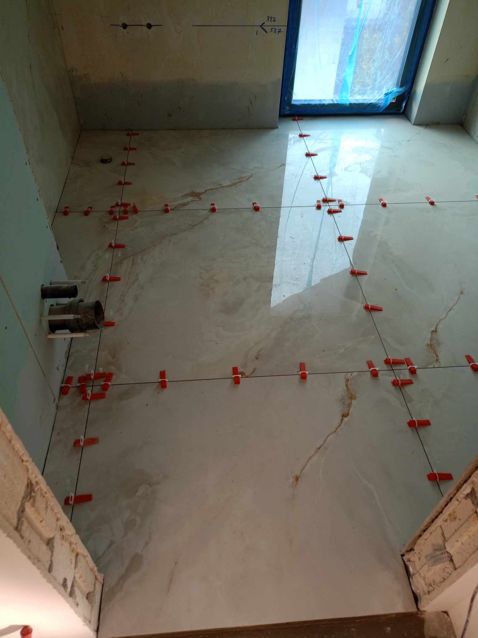 Układanie płytek podłogowych w łazience z użyciem systemu poziomowania, widoczne rury wystające ze ściany i okno zabezpieczone folią.