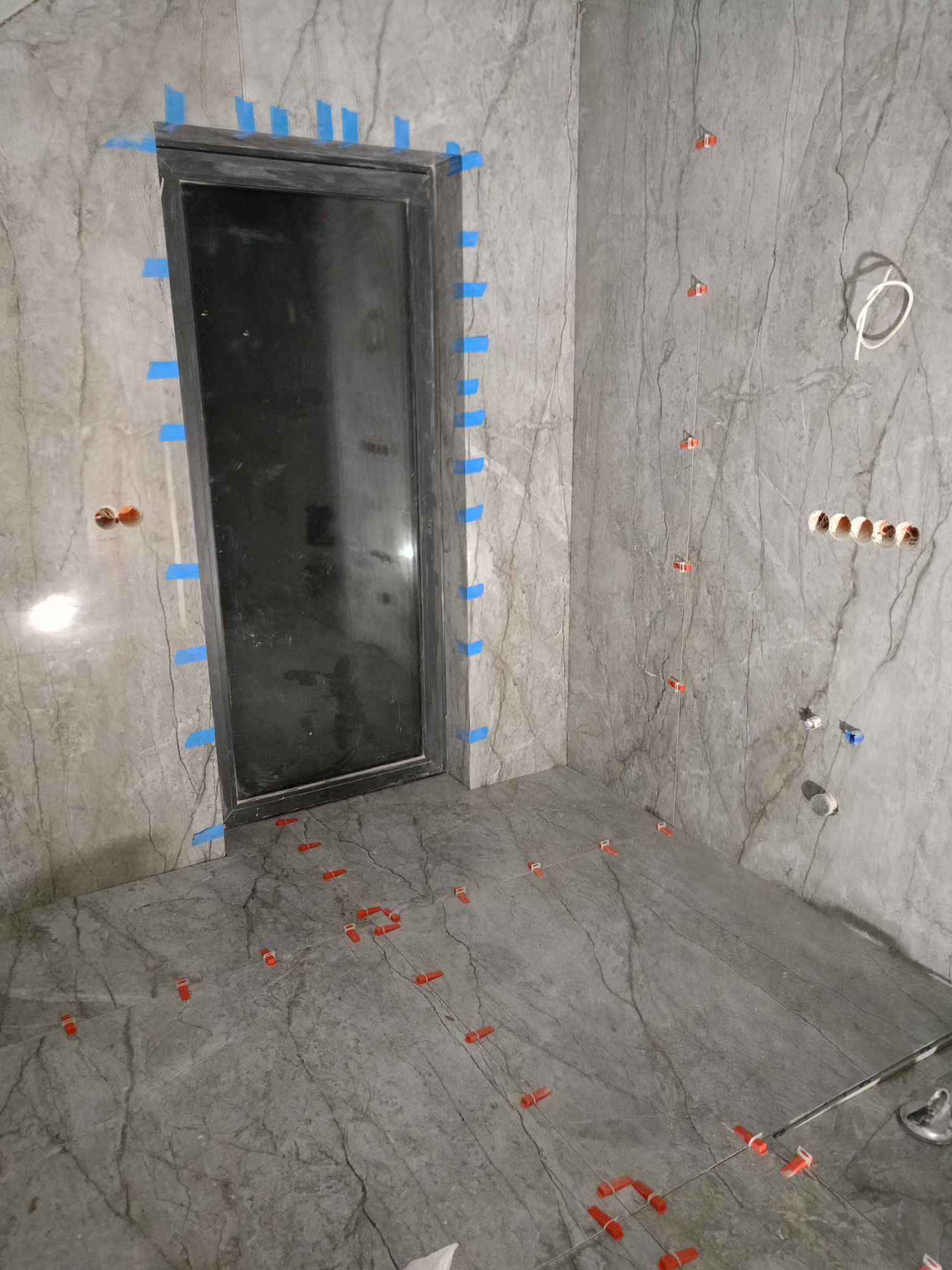 Łazienka w trakcie remontu: szare płytki imitujące marmur na ścianach i podłodze, niebieska taśma malarska wokół czarnej ramy drzwi, system poziomowania płytek z czerwonymi klipsami i żyłkami...