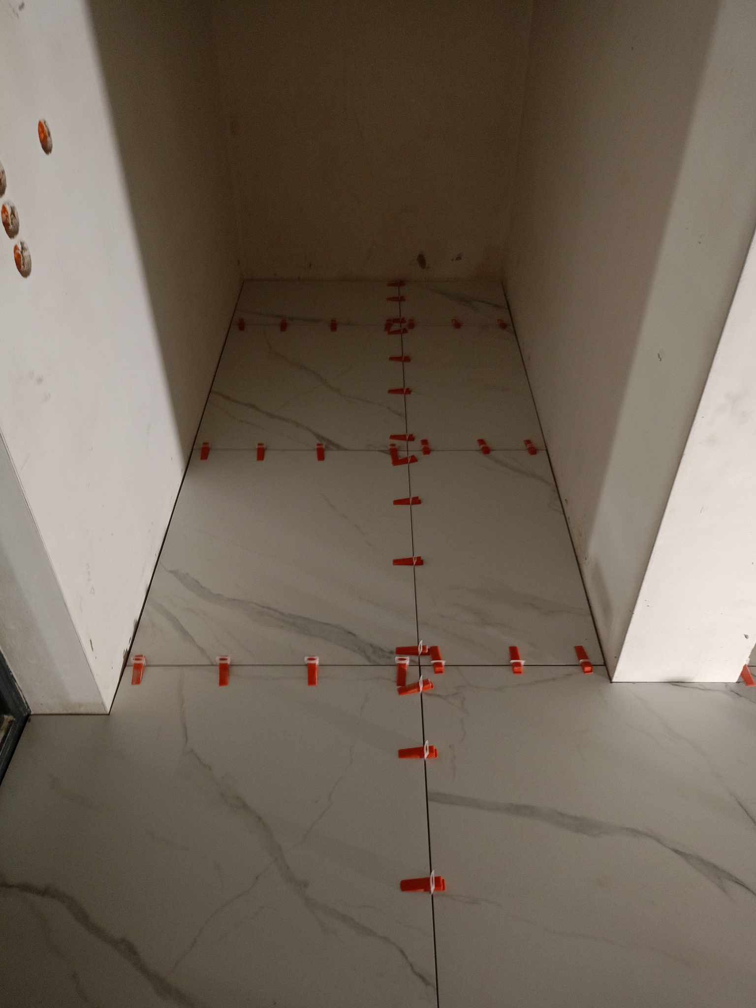 Układanie płytek podłogowych w małej wnęce, z użyciem pomarańczowych klipsów poziomujących, widoczne fugi i wzór marmurkowy płytek.