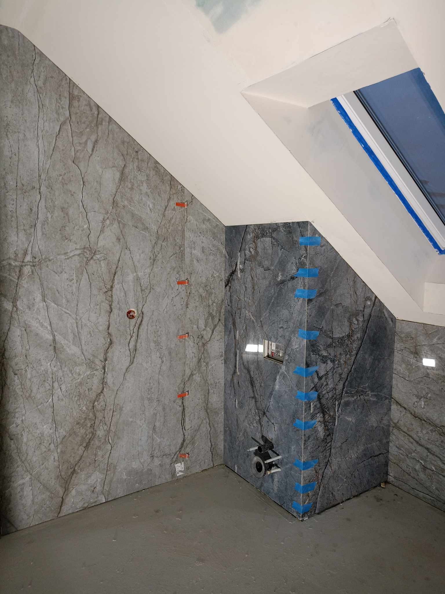 Ściana w łazience wyłożona płytami imitującymi szary marmur z widocznymi elementami instalacji wodnej i elektrycznej, zabezpieczona niebieską taśmą malarską i poziomowana pomarańczowymi klinami...