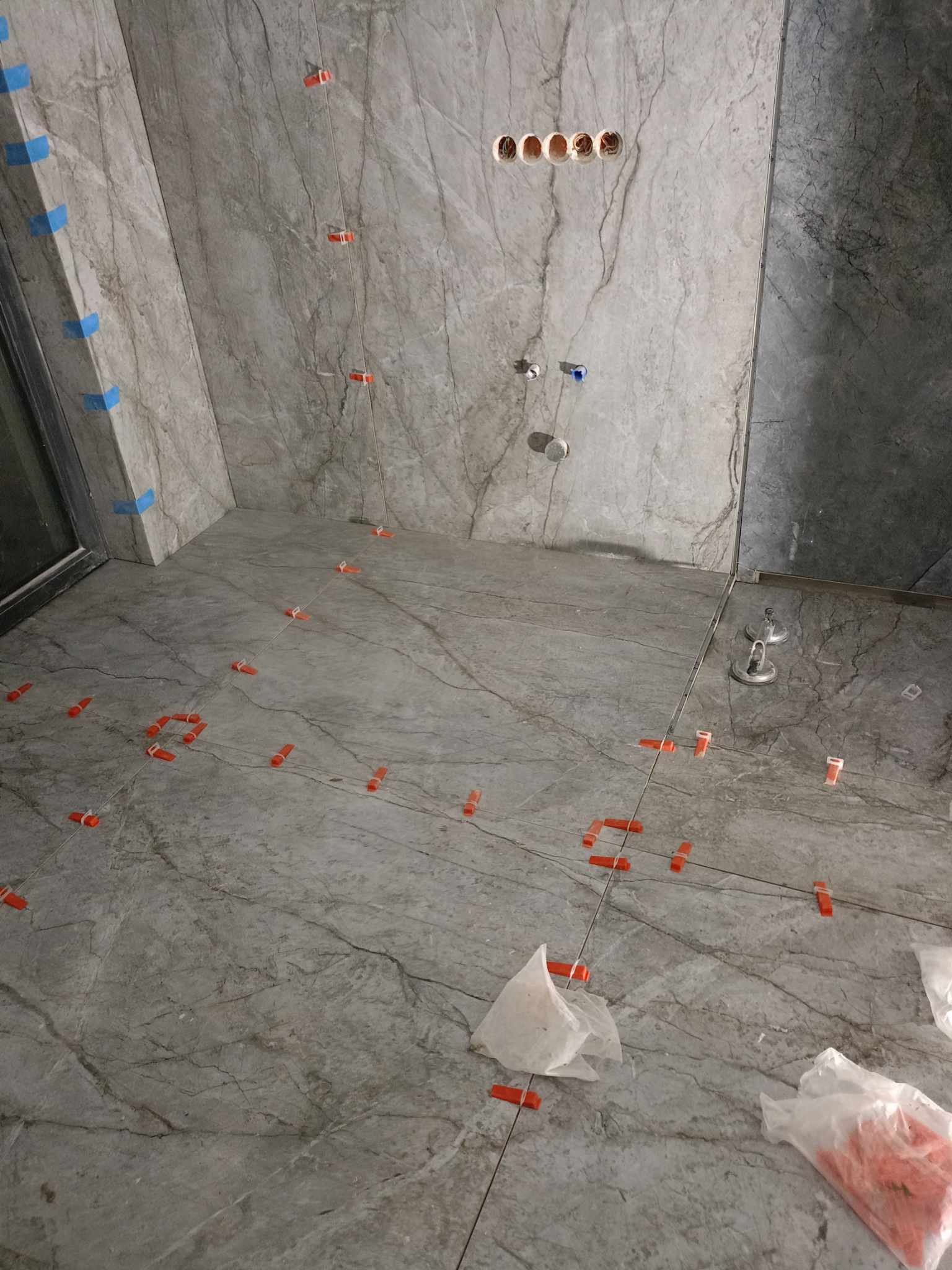 Świeżo ułożone, szare płytki imitujące marmur, z pomarańczowymi krzyżykami dystansowymi i niebieską taśmą malarską na ścianach łazienki w trakcie prac wykończeniowych.