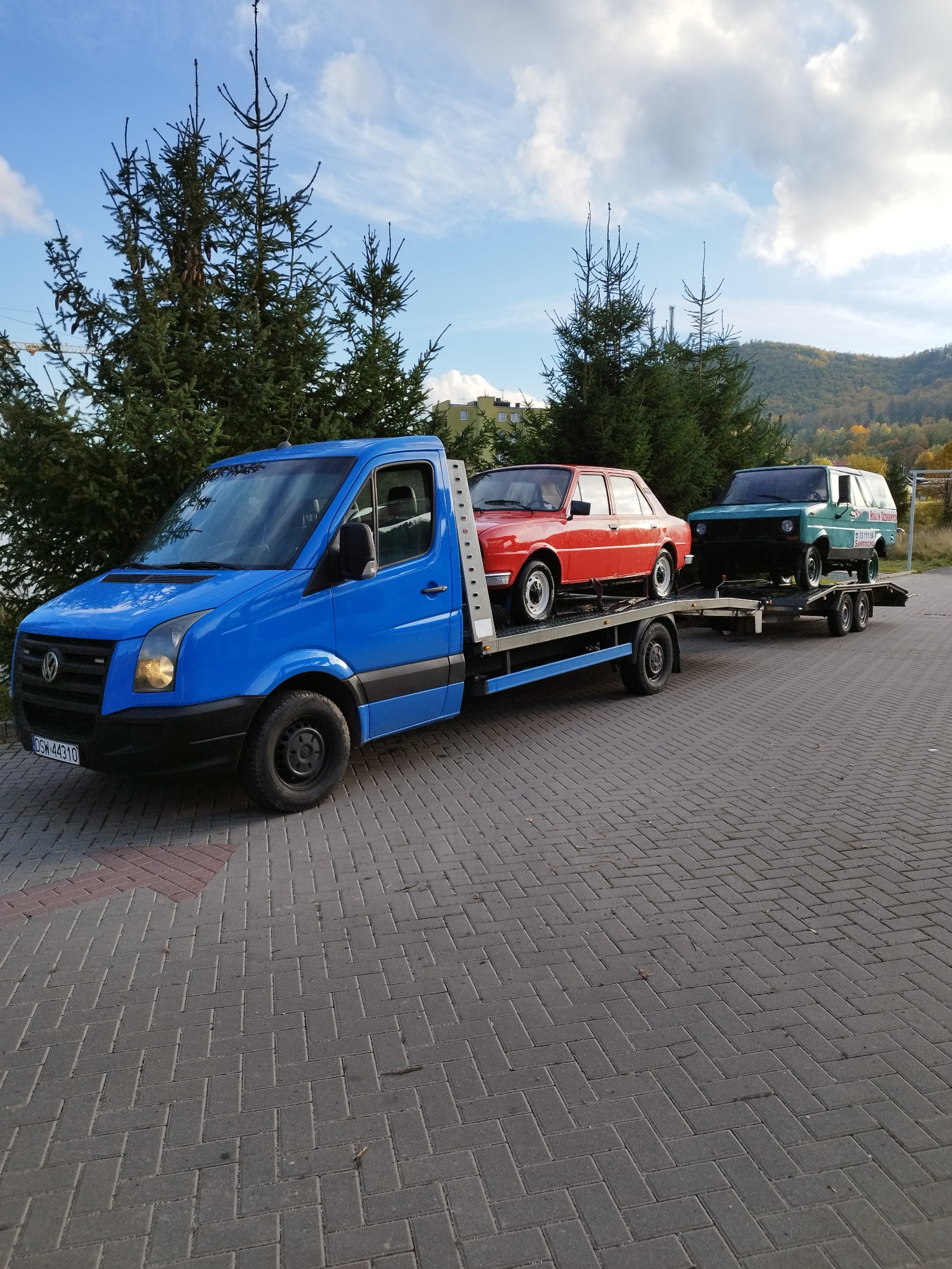 Niebieski samochód transportowy z dwoma starymi samochodami osobowymi na platformie, zaparkowany na brukowanym placu z drzewami iglastymi i wzgórzami w tle.