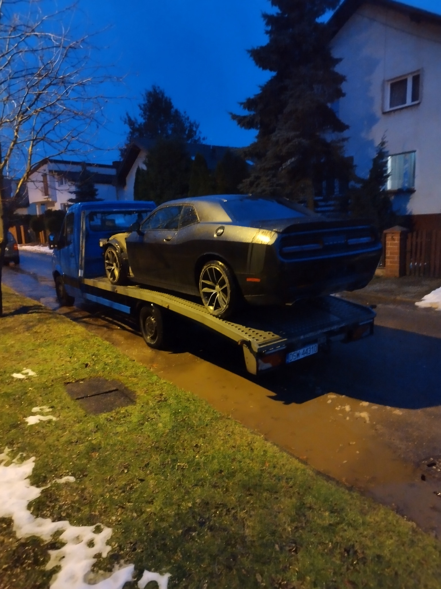 Czarny samochód sportowy Dodge Challenger przewożony na platformie transportowej niebieskiego pojazdu, widok z tyłu, o zmierzchu, z fragmentami śniegu na trawie.