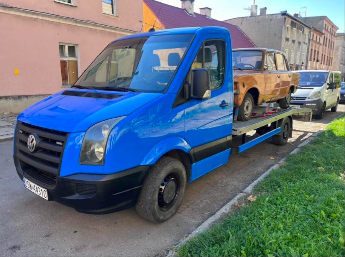 Niebieski Volkswagen Crafter z brązowym samochodem osobowym na platformie, zaparkowany na ulicy z budynkami w tle.
