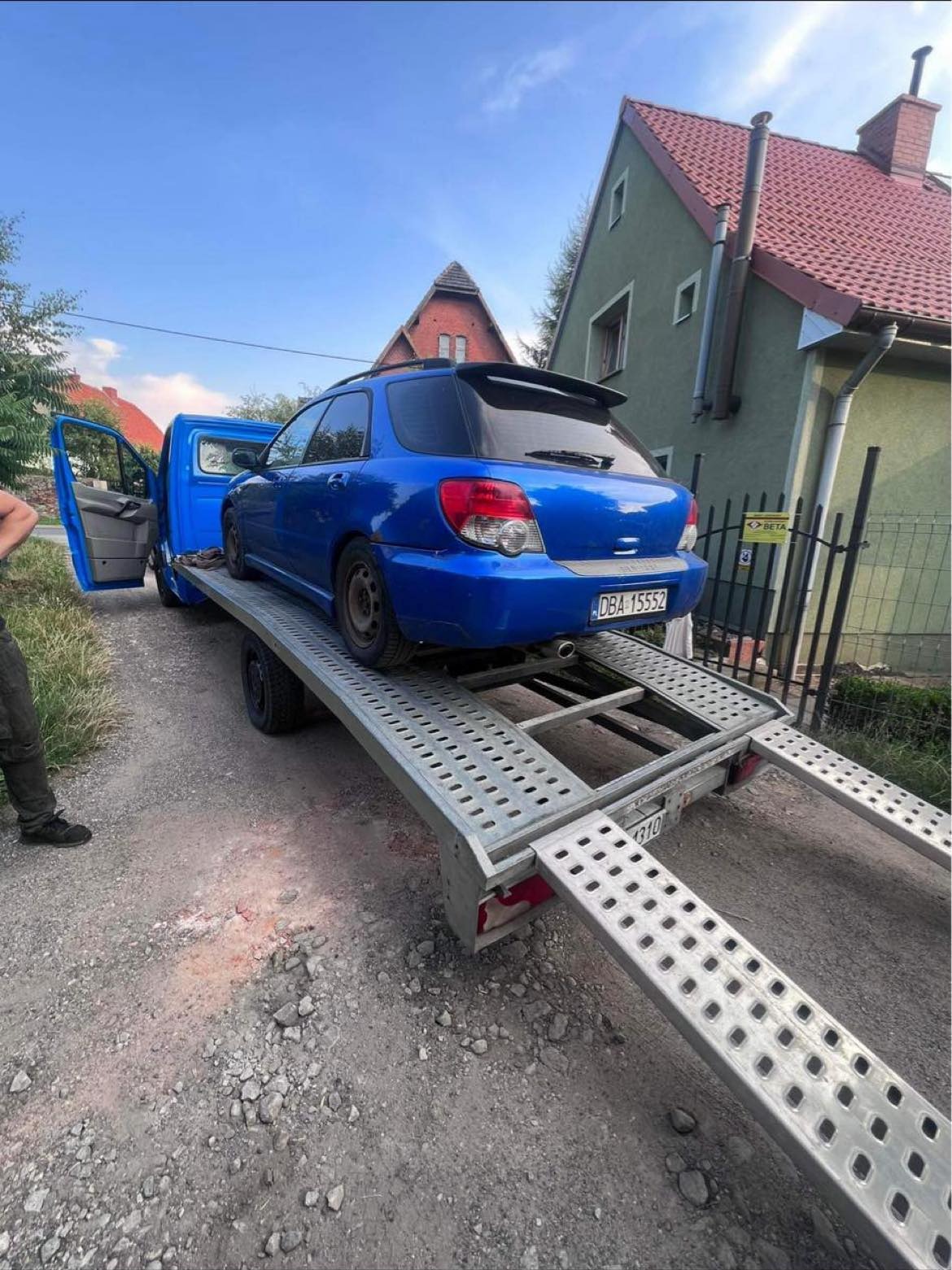 Niebieski samochód kombi Subaru Impreza z widocznym numerem rejestracyjnym DBA 15552 załadowany na srebrną platformę transportową, otwarta kabina niebieskiej ciężarówki i osoba z lewej strony,...