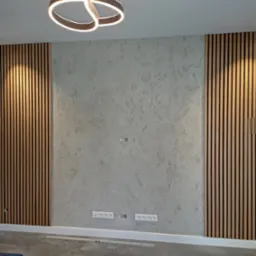 Ściana z dekoracyjnym tynkiem i drewnianymi lamelami po bokach, oświetlona nowoczesną lampą sufitową z okrągłych profili LED, widoczne gniazdka elektryczne i listwa przypodłogowa.