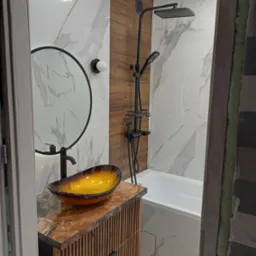 Nowoczesna łazienka w trakcie wykończenia: marmurowe płytki, drewniane panele, czarna armatura, umywalka w odcieniach bursztynu na szafce z lamelami, widoczne niedokończone fragmenty przy drzwiach.