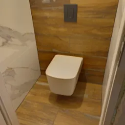 Wisząca toaleta z deską wolnoopadającą na tle drewnopodobnych płytek ściennych i podłogowych, obok ściany wyłożonej płytkami imitującymi marmur.