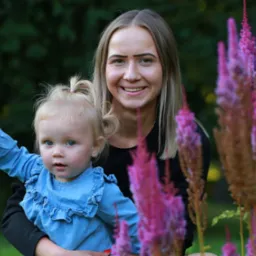 Uśmiechnięta kobieta trzymająca na rękach małą dziewczynkę w dżinsowej sukience na tle zieleni i różowych kwiatów.