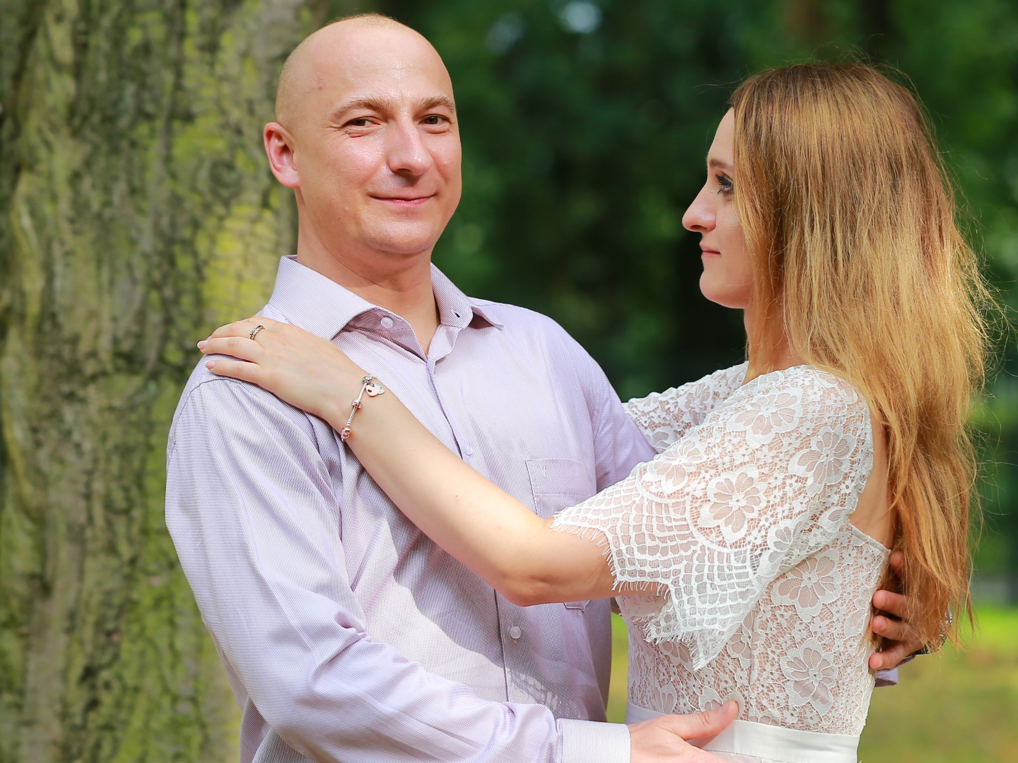 Para obejmująca się na tle zielonego drzewa, kobieta w koronkowej sukience z bransoletką na nadgarstku, mężczyzna w koszuli w paski.