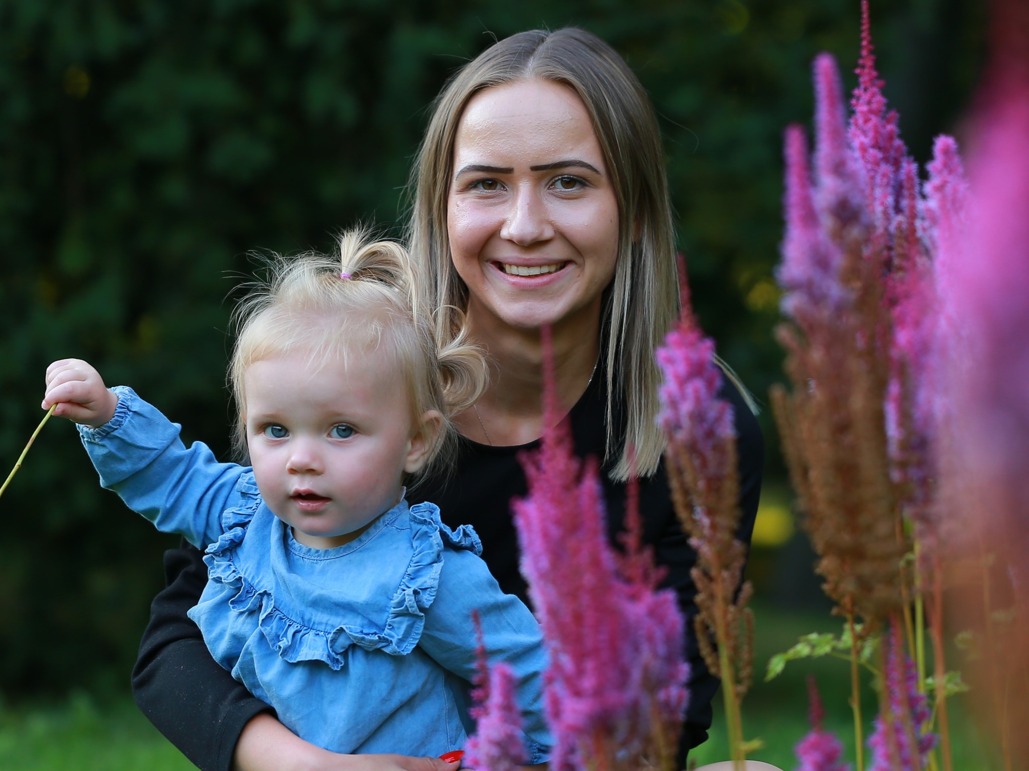 Uśmiechnięta kobieta trzymająca na rękach małą dziewczynkę w dżinsowej sukience na tle zieleni i różowych kwiatów.