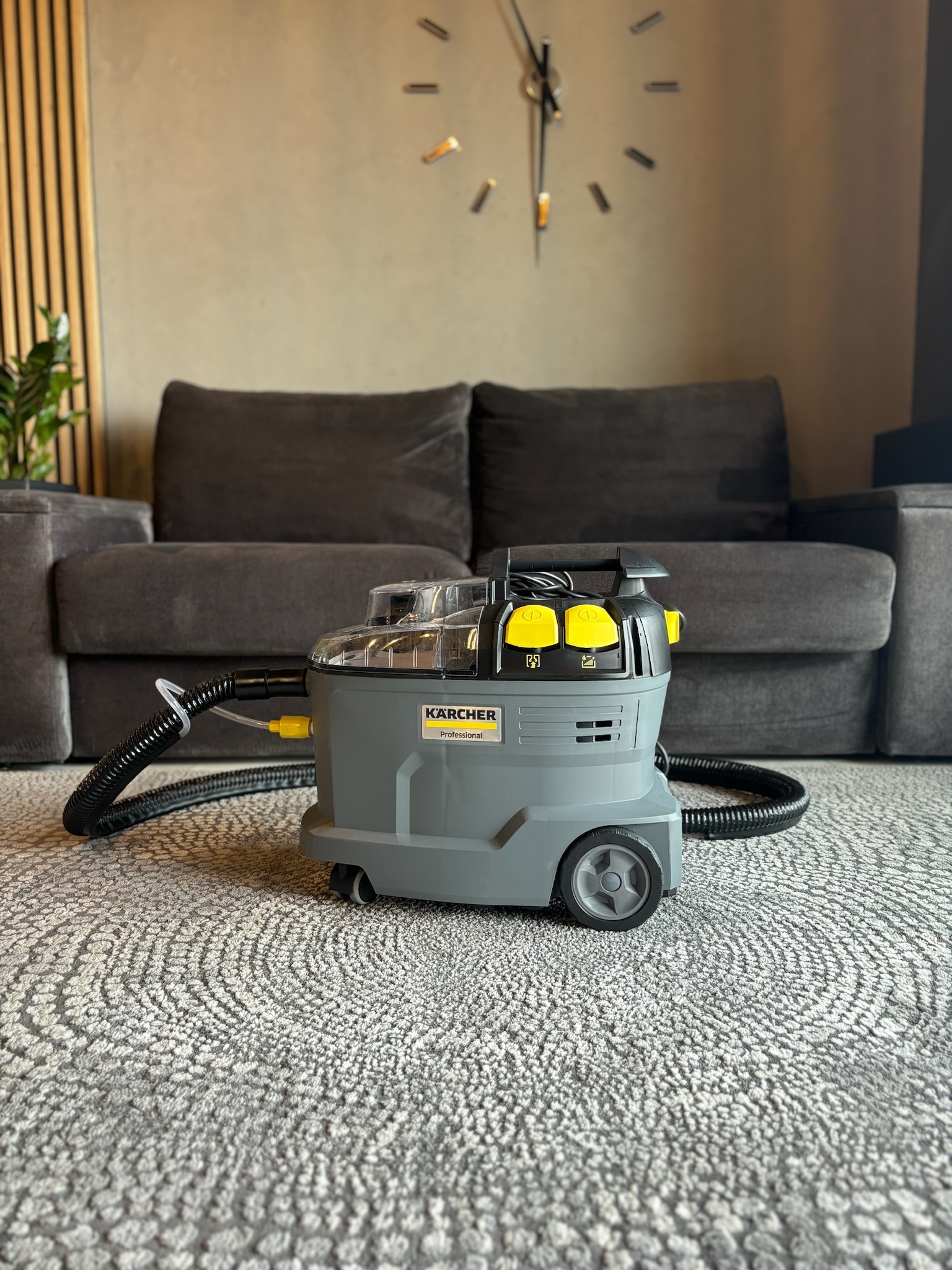 Profesjonalny odkurzacz piorący Kärcher na dywanie ze wzorem, w tle szara kanapa i zegar ścienny.