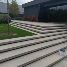 Eleganckie, zewnętrzne schody z jasnych, prostokątnych płyt prowadzące do nowoczesnego budynku z ciemną elewacją i dużymi oknami. Widoczny fragment trawnika i drzewo.