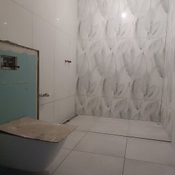 Piter HOME - Łazienka w trakcie wykańczania: białe płytki z motywem liści na ścianach, częściowo zabudowany stelaż WC. Widoczny odpływ liniowy w podłodze.